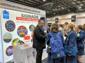 A man in a black jacket stands in front of a sign saying Australian Sign and Graphics Association. He is talking to teenagers in school uniform. 