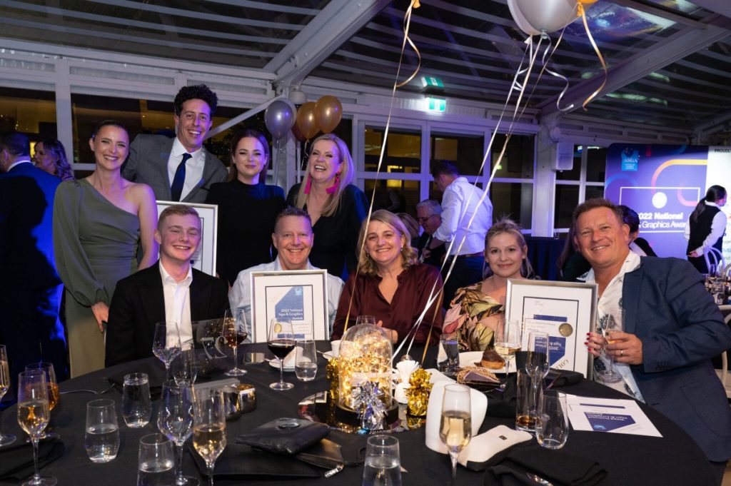 A group of happy people in evening dress sit around a table, some holding awards they have won. There are balloons and gold decorations on the table giving it a festive look. 