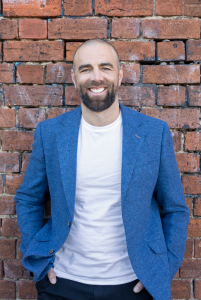 Man with a beard, dressed in white tee shirt and blue sports jacket, leans against a brick wall and smiles.