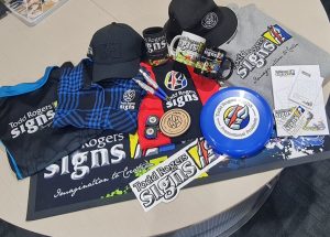 A pile of products from stickers, to tee shirts, a frizbee, mugs and more, all featuring the Todd Rogers Signs logo.
